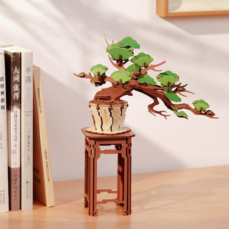 May include: A wooden model of a bonsai tree with green leaves, sitting on a wooden stand. The tree is in a brown pot with a light brown and white pattern.