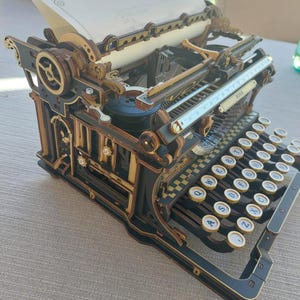 May include: A mechanical typewriter model constructed from layered dark and light wood pieces. It features a QWERTY keyboard with circular keys, a paper carriage with a sheet of paper inserted, and a ROKR brand plate on the front frame.