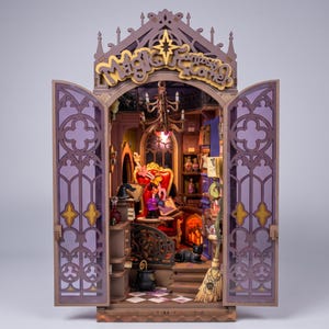 Op de afbeelding: Een miniatuur houten boekenhoek-diorama genaamd Magic Fantasy Corner met open paarse deuren in gotische stijl. Het interieur bevat een rode fauteuil, een kristallen bol, een zwarte kat, een bezem, een kroonluchter en planken vol kleine boeken.