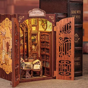 Op de afbeelding: Een houten miniatuur poppenhuis met een boekenkast interieur. Het poppenhuis is bruin geverfd en heeft een bord met de tekst "Boekhandel Herinneringen". Het poppenhuis is open en het interieur is zichtbaar. De boekenkast is gevuld met boeken en er zijn andere decoratieve items in het poppenhuis.
