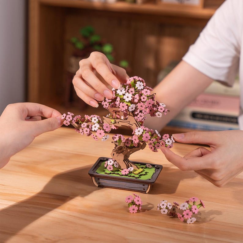May include: A wooden 3D puzzle of a cherry blossom bonsai tree with pink and white flowers. The tree is assembled and sitting on a wooden table. There are extra flowers on the table.