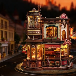 May include: Miniature model of a European-style street scene at dusk. The detailed diorama features a multi-story building with a cafe, illuminated windows, and a sign that reads "Fresh Taste". The street is lined with buildings, trees, and flower pots.