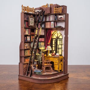 Puede incluir: Un kit de rincón de lectura en miniatura de madera con una estantería de esquina llena de libros diminutos, una escalera negra alta, un escritorio amarillo con silla, una ventana arqueada iluminada y una pequeña lámpara de madera cortada a láser.