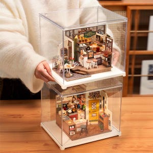 May include: Two miniature dioramas of a coffee shop and a kitchen, each housed in a clear acrylic display case. The coffee shop has a sign saying "Coffee Shop". The kitchen features a yellow door. Both are filled with tiny details.