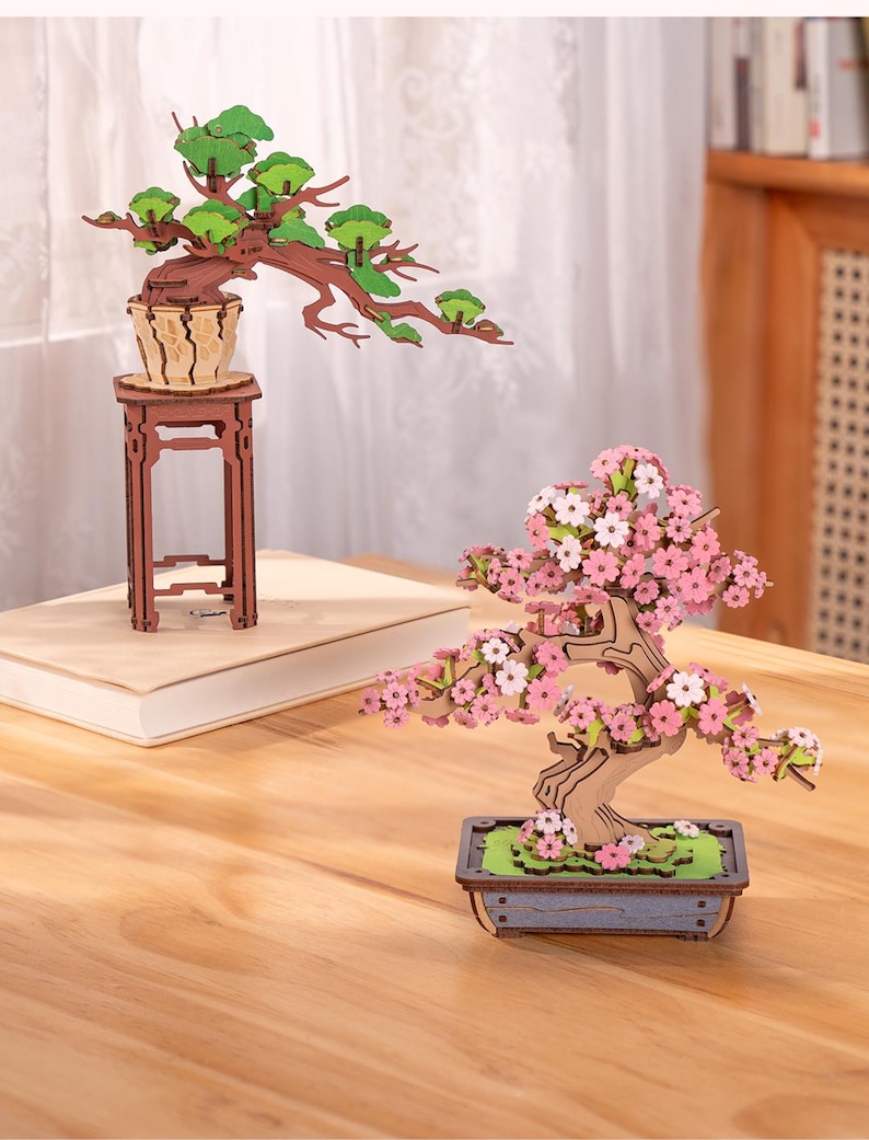 May include: Two wooden bonsai trees, one with green leaves and the other with pink flowers. Both trees are on wooden stands and are in pots.