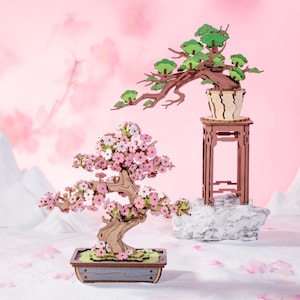 May include: Two wooden bonsai trees, one with pink blossoms and the other with green leaves, are displayed on a white surface. The bonsai with pink blossoms is in a brown pot, while the bonsai with green leaves is in a brown pot and on a brown stand.