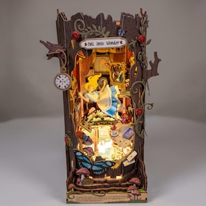 Puede incluir: Un diorama de madera para estantería que muestra a un personaje con vestido azul cayendo a través de un tronco de árbol. La estructura incluye un reloj de bolsillo, rosas rojas, mariposas, setas y un letrero que dice Fall into Wonder en cursiva.