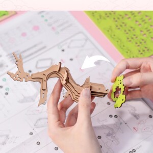 May include: A person assembling a wooden model of a tree with detailed instructions. The model is made of brown wood and has a green leaf design. The person is holding the model in their left hand and a green leaf piece in their right hand.