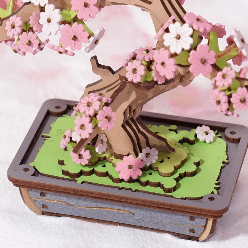 May include: A wooden model of a cherry blossom bonsai tree with pink and white flowers. The tree is in a brown wooden pot with a gray base.