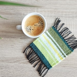 May include: A white coffee mug with a black handle filled with coffee sits on a green and white striped woven coaster with black fringe.