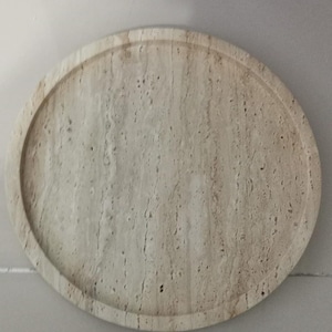 Puede incluir: Una bandeja circular hecha de piedra travertino beige, con una textura porosa natural y un borde elevado. La superficie muestra vetas lineales caracter&iacute;sticas y peque&ntilde;os orificios en el material de color tostado claro, creando un patr&oacute;n org&aacute;nico.