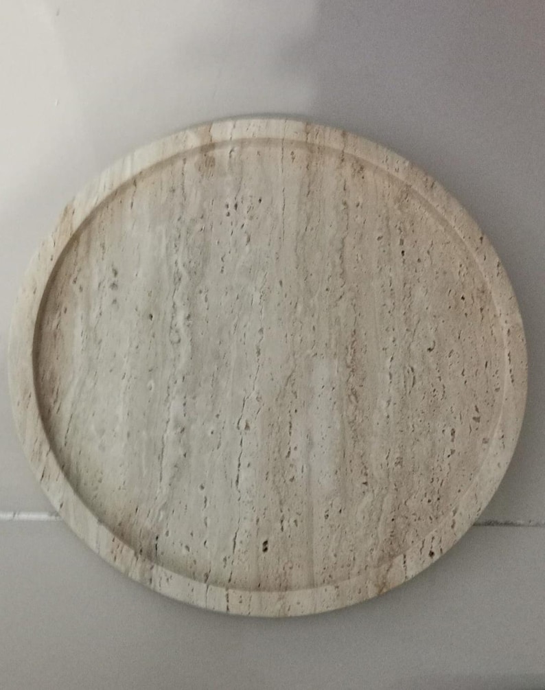 Puede incluir: Una bandeja circular hecha de piedra travertino beige, con una textura porosa natural y un borde elevado. La superficie muestra vetas lineales caracter&iacute;sticas y peque&ntilde;os orificios en el material de color tostado claro, creando un patr&oacute;n org&aacute;nico.