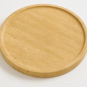 Puede incluir: Una bandeja circular hecha de piedra travertino de color beige claro, con una superficie lisa y plana, un borde exterior elevado y una textura natural porosa caracter&iacute;stica de la piedra, mostrando sutiles variaciones de color y veteado.