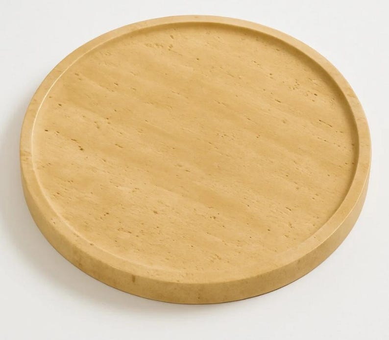 Puede incluir: Una bandeja circular hecha de piedra travertino de color beige claro, con una superficie lisa y plana, un borde exterior elevado y una textura natural porosa caracter&iacute;stica de la piedra, mostrando sutiles variaciones de color y veteado.