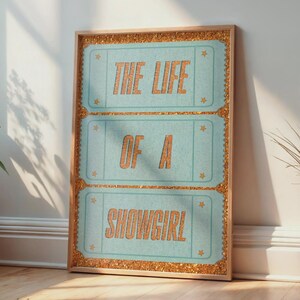 Può includere: Un poster incorniciato con la scritta "THE LIFE OF A SHOWGIRL" in lettere arancioni su sfondo azzurro. La cornice è di colore marrone chiaro con bordi glitterati dorati. Il poster è appoggiato a un muro bianco.