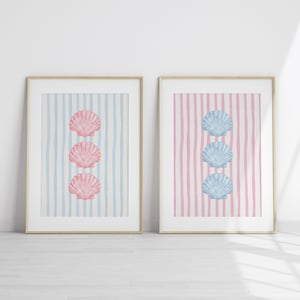 May include: Two framed art prints featuring seashell designs. One print has three pink seashells on a blue and white striped background. The other print has three blue seashells on a pink and white striped background.