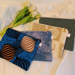 Peut inclure: Cinq pochettes zippées en tissu avec différents motifs. Les pochettes sont disposées sur un fond en tissu blanc. Les motifs comprennent un design géométrique bleu et orange, un motif tie-dye bleu et blanc, un motif floral beige et blanc, un motif d'oiseau beige et blanc et un motif géométrique noir et blanc.