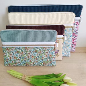 May include: A collection of four rectangular zippered pouches arranged in a row. Each pouch features a top section of solid-colored corduroy fabric and a bottom section of floral-patterned fabric, separated by a white zipper and trim.