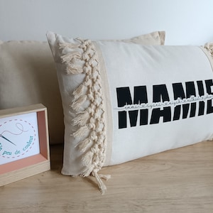 Peut inclure: Un oreiller blanc avec un motif noir "MAMIE" et une bordure à franges. L'oreiller est sur une surface en bois avec une petite boîte en bois qui dit "Un peu de couleur" dans un cercle.