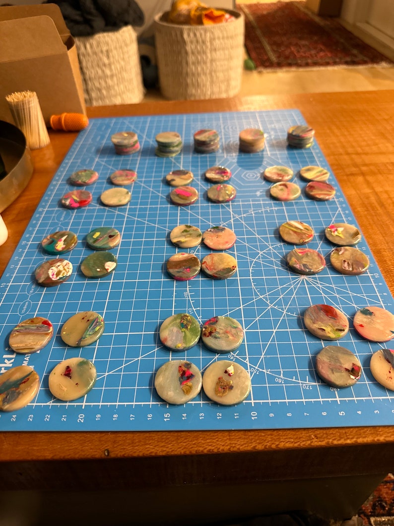 May include: A blue cutting mat with 25 round, colourful, resin coasters. The coasters are arranged in rows and columns on the mat. The coasters are made of clear resin with embedded colourful flakes and swirls.