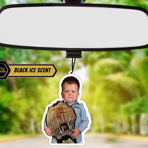 May include: A car air freshener featuring a young boy wearing a red hat and holding a gold championship belt. The air freshener is white with black text that reads "BLACK ICE SCENT".