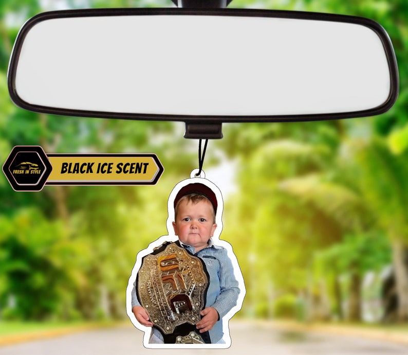 May include: A car air freshener featuring a young boy wearing a red hat and holding a gold championship belt. The air freshener is white with black text that reads "BLACK ICE SCENT".