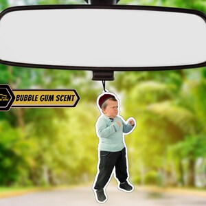 May include: A car air freshener featuring a cartoon image of a young boy wearing a red hat and a blue shirt. The air freshener is shaped like a circle and has the text "BUBBLE GUM SCENT" printed on it.