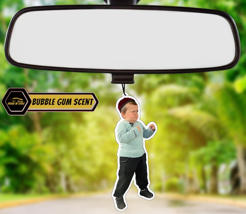 May include: A car air freshener featuring a cartoon image of a young boy wearing a red hat and a blue shirt. The air freshener is shaped like a circle and has the text "BUBBLE GUM SCENT" printed on it.