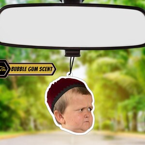 May include: A car air freshener with a picture of a young boy wearing a red hat. The air freshener is shaped like a circle and has the text "BUBBLE GUM SCENT" on it.