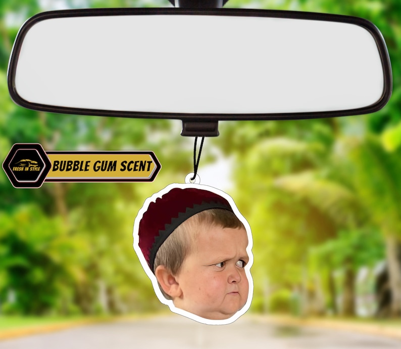 May include: A car air freshener with a picture of a young boy wearing a red hat. The air freshener is shaped like a circle and has the text "BUBBLE GUM SCENT" on it.