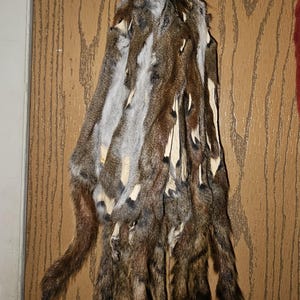 May include: A collection of multiple brown and grey animal fur pelts hanging from a metal ring. Each pelt features a mix of dark brown, light brown, and grey fur with visible light-colored leather backing along the edges of the strips.
