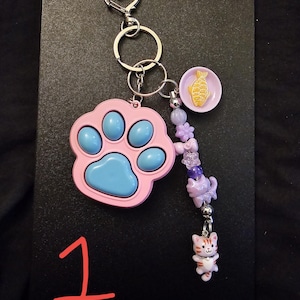 May include: A keychain featuring a pink and blue paw-shaped fidget toy, a small purple plate with a yellow fish charm, and a beaded strand with purple beads, a cat-shaped bead, and a small orange and white striped cat charm with a heart clasp.