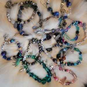 May include: A collection of beaded bracelets in various colors, including blue, green, and pink, with charms and letter beads spelling out words like "WOLF" and "AWOL". The bracelets are displayed on a white, fluffy surface.