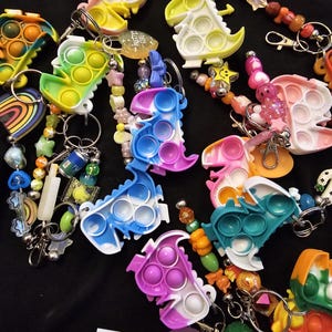 May include: A collection of dinosaur-shaped silicone sensory keychains in various tie-dye colors, each featuring circular push-pop bubbles. The keychains are attached to metal rings with assorted colorful plastic beads, charms, and small trinkets.