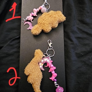 May include: Two keychains featuring fuzzy brown dinosaur-shaped plush charms. Each keychain includes a silver-tone metal clasp, a pink crescent moon charm, and a strand of pink beads with small dinosaur figures. One keychain also features a lollipop charm.