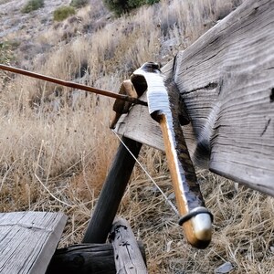 May include: A wooden recurve bow with a dark, polished finish and a silver-coloured tip, resting against a weathered wooden plank. An arrow with white fletching is positioned horizontally across the bow, pointing towards the left side of the frame.