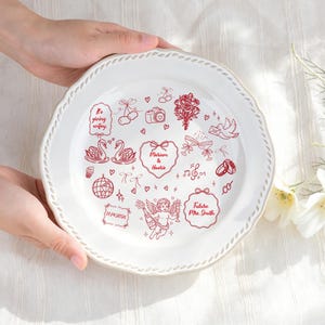 Könnte beinhalten: Ein weißer Keramikt weißer Keramikteller mit gewelltem Rand und roten Strichzeichnungen von Schwänen, einem Blumenstrauß, einer Kamera, einer Discokugel, einem Putto sowie den Texten "Mariam & Haarie", "11.14.2026", "Future Mrs. Smith" und "is giving wifey."