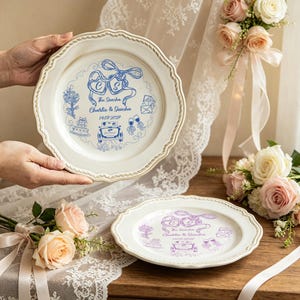 Puede incluir: Dos platos decorativos blanquecinos con borde festoneado y borde tipo cuerda. Los platos presentan un diseño de boda azul y morado con los nombres "Charlie & Sascha" y la fecha "14.07.2027".