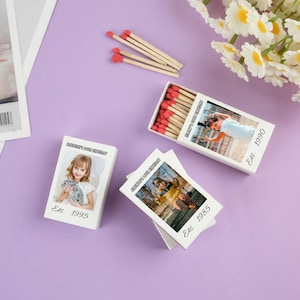 Peut inclure: Des boîtes d'allumettes blanches avec des photos et du texte, dont "MOMENT'S FOR BIRTHDAY" et des dates comme "Est. 1995" et "Est. 1990". Les allumettes ont des têtes rouges. Quelques allumettes en vrac sont éparpillées à proximité. Le fond est violet clair.