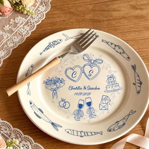 May include: A white ceramic plate decorated with blue illustrations of fish, hearts, and wedding symbols. The plate is personalised with the names "Charlie & Sascha" and the date "14.07.2027". A silver fork with a wooden handle is placed on the plate.