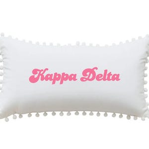 May include: White rectangular cushion featuring the pink text "Kappa Delta." The cushion is trimmed with small pom-poms. A decorative cushion suitable for a bedroom or living space.