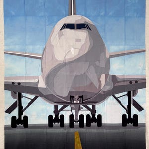 May include: A large, gray airplane quilt with a blue sky background. The quilt depicts the front of a commercial jet on a runway, with the nose and cockpit prominently displayed. The landing gear and a yellow runway stripe are visible.