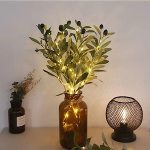 Könnte beinhalten: Eine braune Glasvase mit einem um den Hals gewickelten Band, gefüllt mit einem grünen Olivenzweig mit kleinen schwarzen Oliven und weißen Lichterketten.