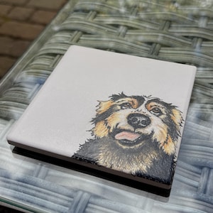 May include: Square ceramic coaster with a detailed illustration of a dog's face. The dog has black, brown, and tan fur, with its tongue sticking out. The coaster has a white surface and a dark brown edge.