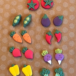 May include: A collection of colorful, miniature vegetable-shaped embellishments. The assortment includes tomatoes, peas, eggplants, carrots, peppers, radishes, beets, and broccoli. Each piece is brightly colored and detailed.