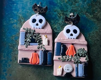 May include: A pair of dangle earrings featuring a pink arched bookshelf design. Each earring displays a white skull at the top, a black crescent moon, books, a green trailing plant, a bright orange pumpkin, and a small lit candle or crystal ball.