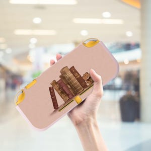 Può includere: Una custodia rosa e gialla per console di gioco portatile con un'illustrazione di libri antichi e un piccolo orsacchiotto. La custodia ha uno sfondo rosa chiaro con una fila di libri marroni e un piccolo orsacchiotto.