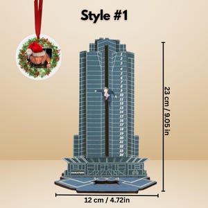 May include: A blue and gray skyscraper-shaped ornament with a figure hanging from it. The ornament is 9.05 inches tall and 4.72 inches wide. A smaller round ornament with a Santa hat is attached. The text "NAKATOMI" is visible.