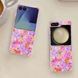 May include: Two open flip phones with floral designs. The phones have a vibrant pattern of pink, purple, and orange flowers. The screens display a blue and green gradient and a circular image. The phones are on a light beige surface.