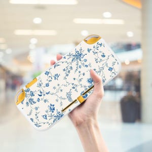Könnte beinhalten: Eine cremefarbene Hülle für eine Handheld-Spielkonsole mit blauem Blumenmuster. Das Gehäuse hat ein blaues Blumenmuster auf cremefarbenem Grund. Die Seiten und Knöpfe sind gelb. Für eine Handheld-Spielkonsole.